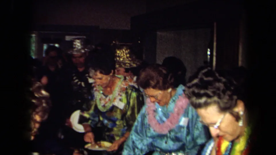 1971 California home-movie frame with adults in leis and patterned clothing serving food at a buffet in a dim private interior.