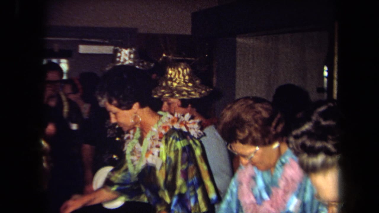 1971 California home-movie frame showing adult guests in floral leis and metallic hats at a crowded indoor party.