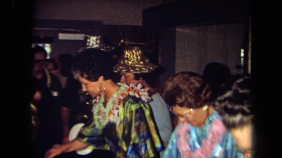 1971 California home-movie frame showing adult guests in floral leis and metallic hats at a crowded indoor party.