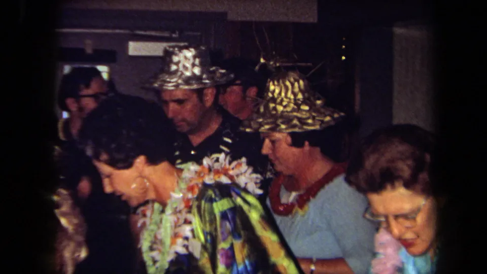 1971 California frame with guests wearing floral leis, metallic hats, glasses, and colorful clothing during an indoor party.