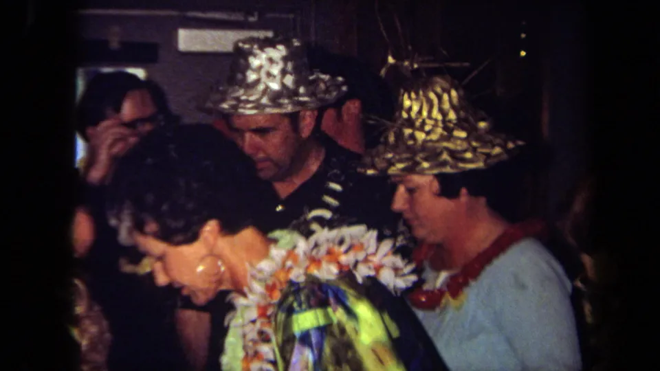 1971 California party footage frame with a woman in a floral lei near adults in metallic hats and casual clothing.