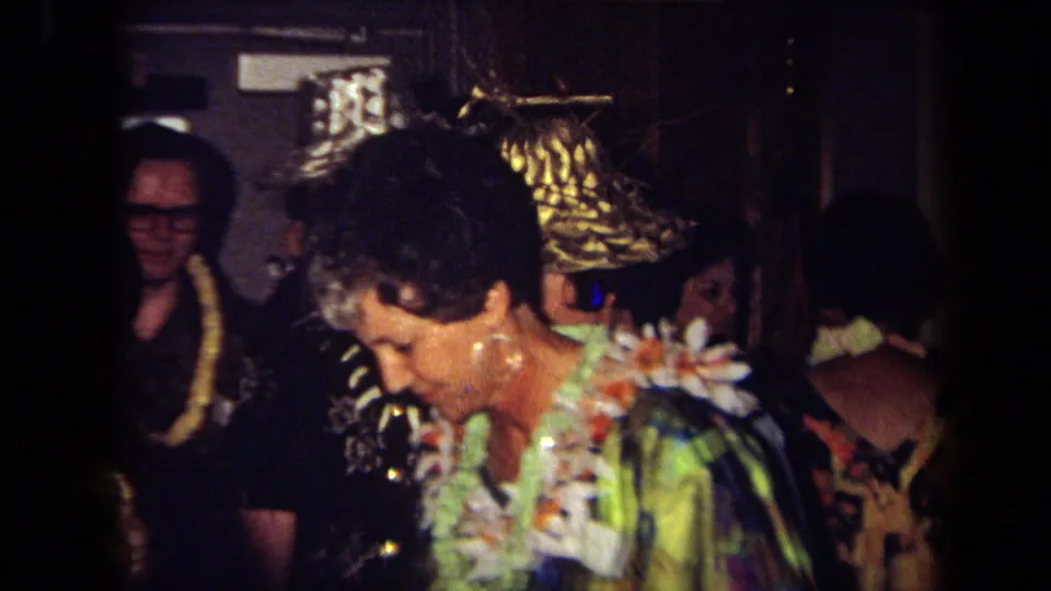 1971 California home-movie frame showing a woman in a floral lei and colorful garment beside other adult party guests.