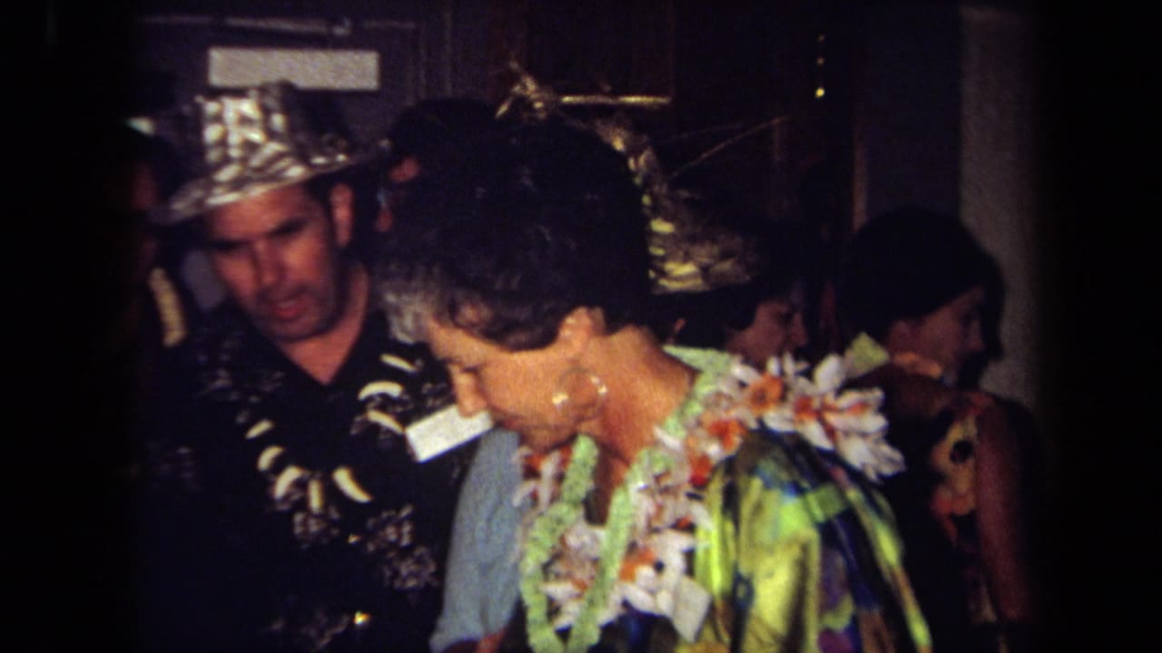 1971 California home-movie frame showing adults in leis and tropical clothing inside a crowded party room.
