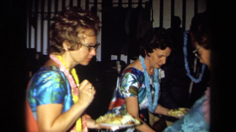 Women at a 1971 California Hawaiian-themed party hold food plates while standing in a warmly lit interior.