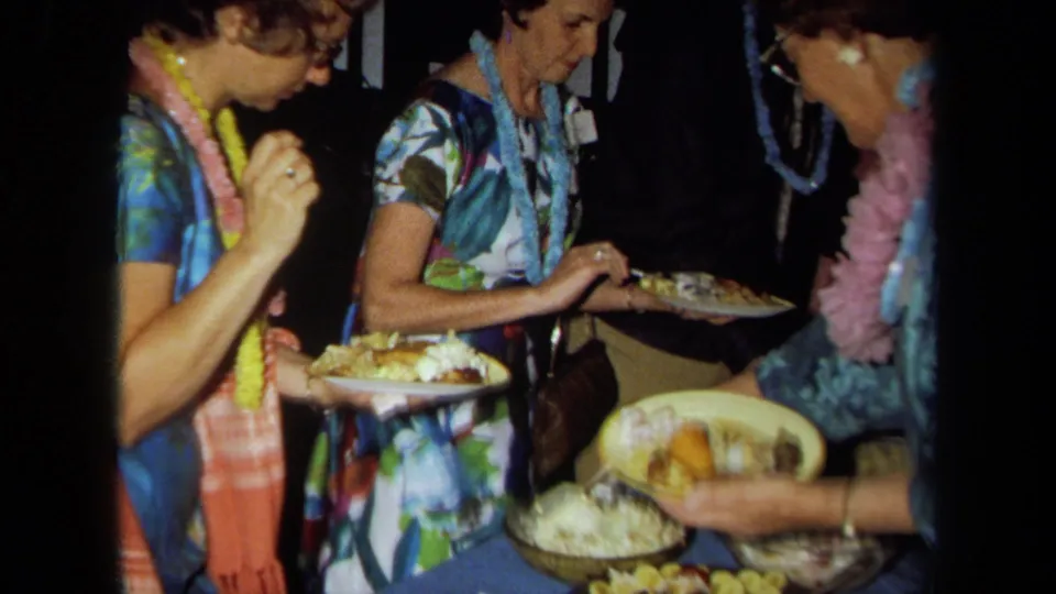 Adults in leis and patterned dresses gather around food plates in a crowded 1971 California party interior.