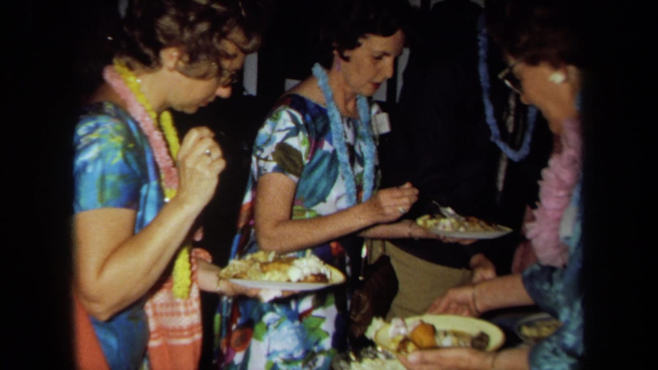 1971 California home-movie frame of women in tropical dresses and flower leis serving food at a Hawaiian-themed party.