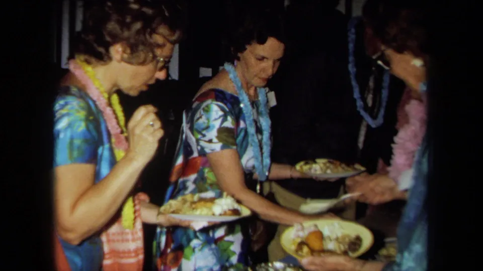 Women wearing flower leis and colorful dresses serve themselves from a potluck table in 1971 California home-movie footage.