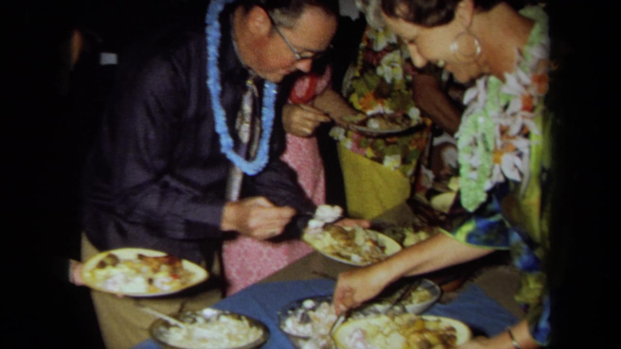 Adults wearing leis serve food from a buffet table at a dimly lit party.