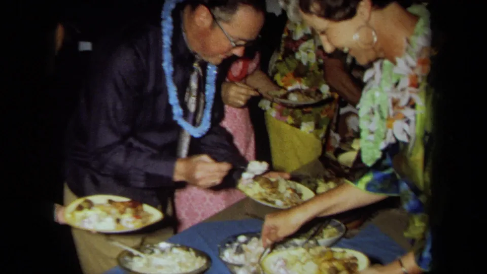 1971 California home-movie frame showing adults in leis and tropical clothing serving buffet food at a luau-style gathering.