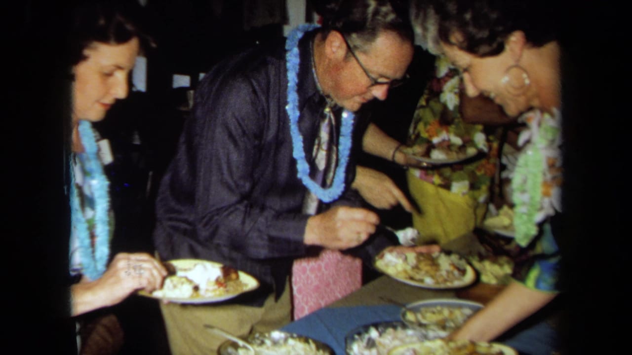 1971 California home-movie frame with a man in glasses and a blue lei serving food beside guests in colorful luau attire.