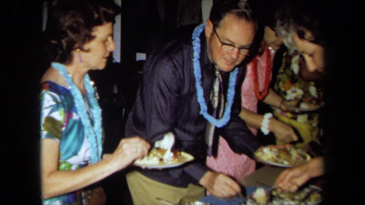 1971 California home-movie frame showing adults in leis and tropical clothing holding plates around a buffet table.