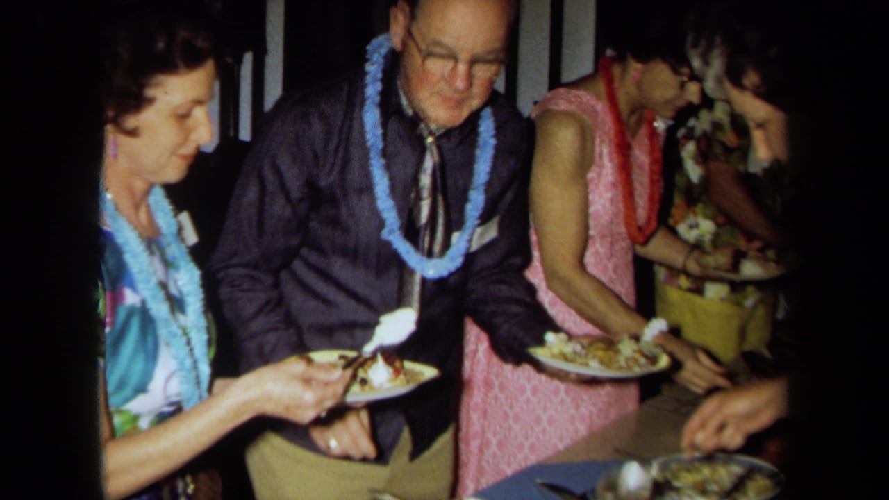 1971 California home-movie frame of adults wearing leis and bright clothing serving themselves at a luau-style buffet.