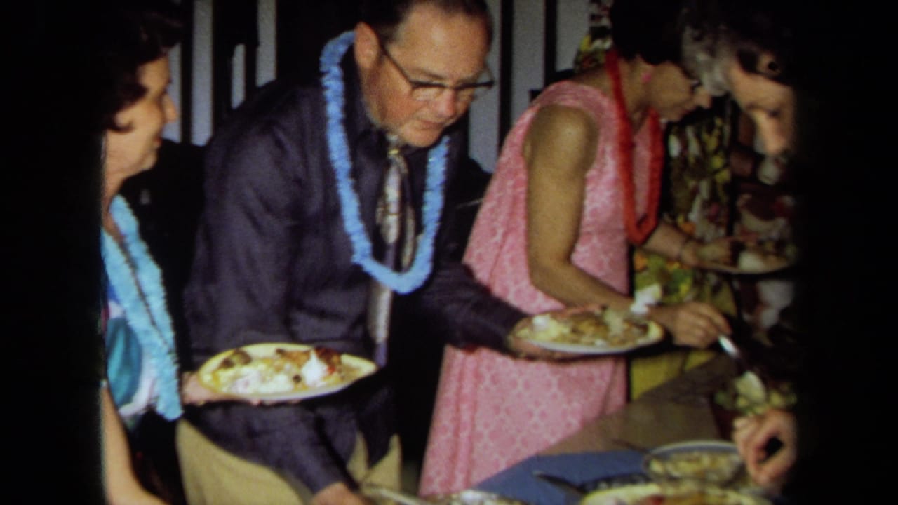 1971 California home-movie frame of adults in leis and bright clothing serving food at an indoor party buffet.