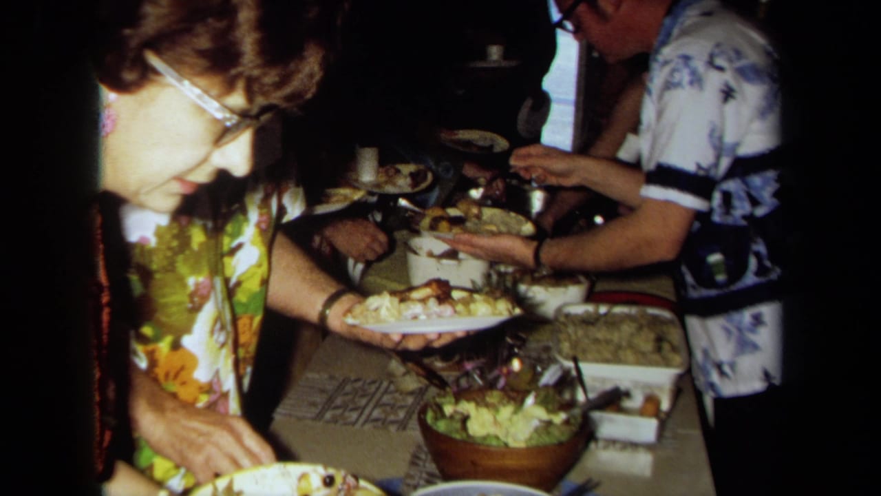 A woman in glasses serves food from a crowded buffet table as other guests stand nearby.