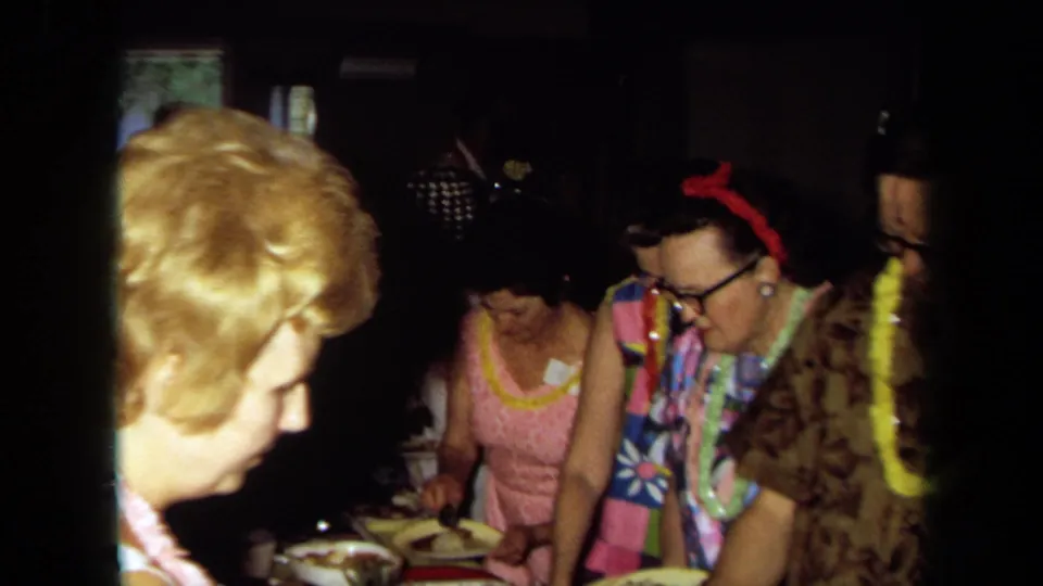 Adults at a 1971 California Hawaiian-themed party stand around buffet dishes, wearing leis, colorful clothing, and eyeglasses.