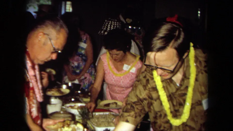 1971 California home-movie frame of adults in leis and patterned clothing serving themselves from a crowded indoor buffet.