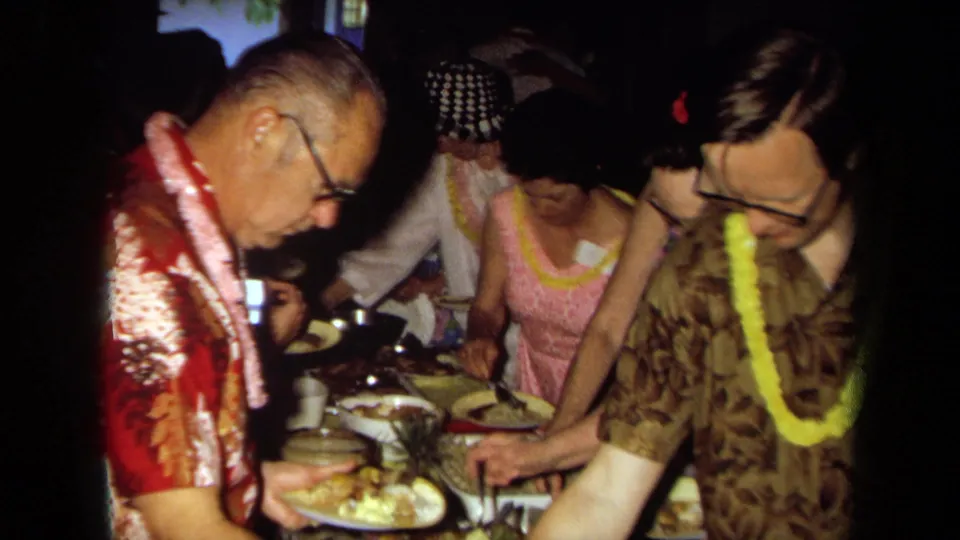 Adults at a 1971 California Hawaiian-themed indoor party serve food from a buffet while wearing leis and patterned shirts.