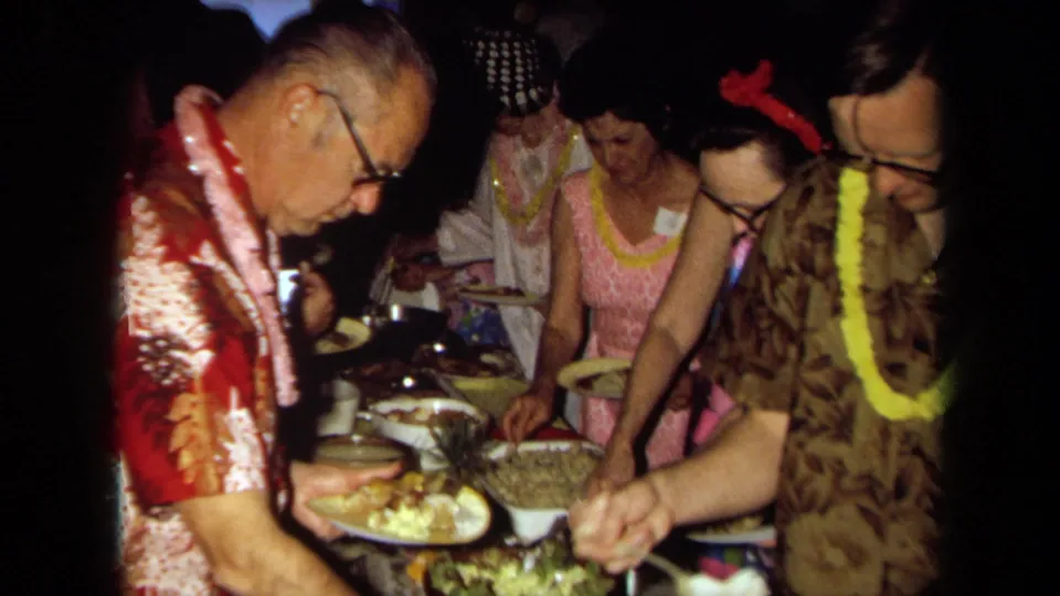 1971 California home-movie frame of adults wearing leis and patterned clothing serving food from a buffet table.