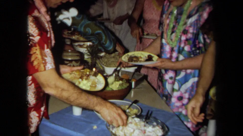 Adults in colorful 1970s clothing stand close together at a California buffet table, holding plates and serving food.