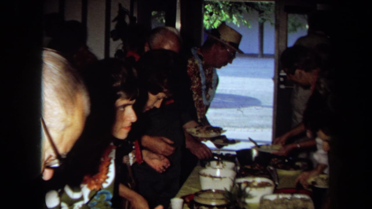 1971 California home-movie frame showing adults serving food at a long table, with leis, pots, plates, and bright outdoor light beyond.
