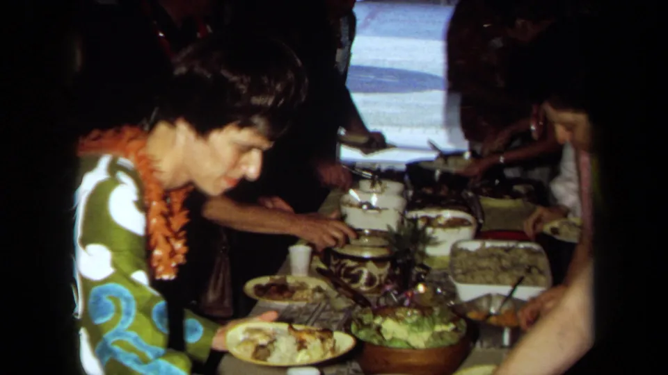 1971 California frame with a woman in patterned clothing and a lei beside plates, salad, and serving dishes at a communal meal.