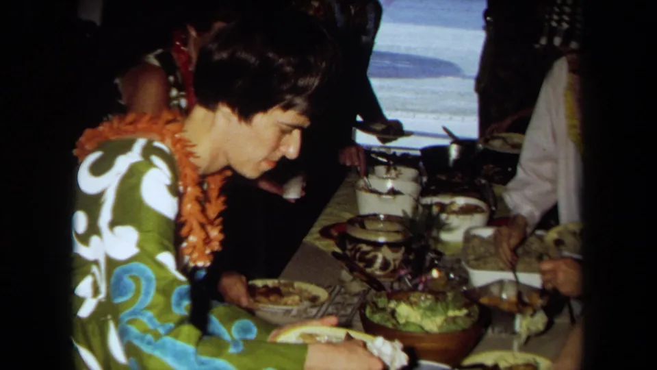 Home-movie frame from 1971 California showing a guest in a lei with a plate near bowls, pots, and other adults at a shared meal.