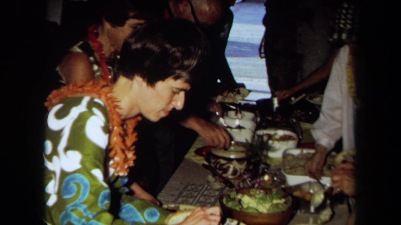 1971 California home-movie frame of a woman wearing a lei beside guests and serving dishes at a Hawaiian-themed party buffet.