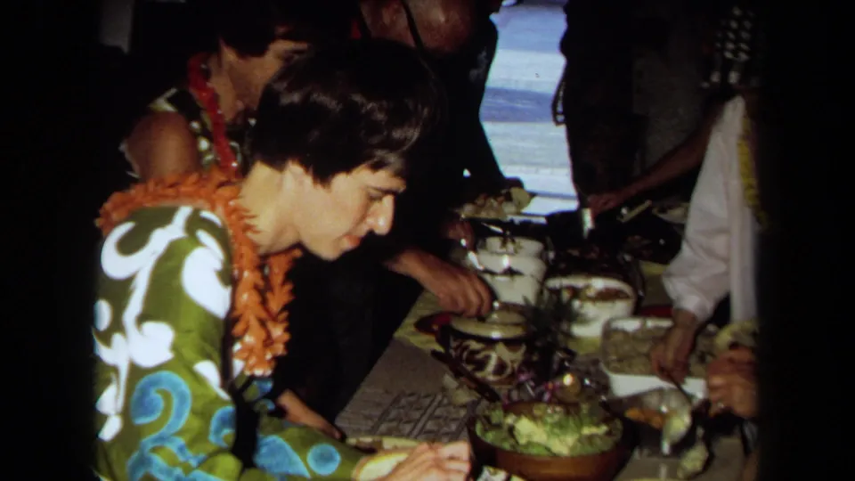 1971 California home-movie frame of a woman wearing a lei beside guests and serving dishes at a Hawaiian-themed party buffet.
