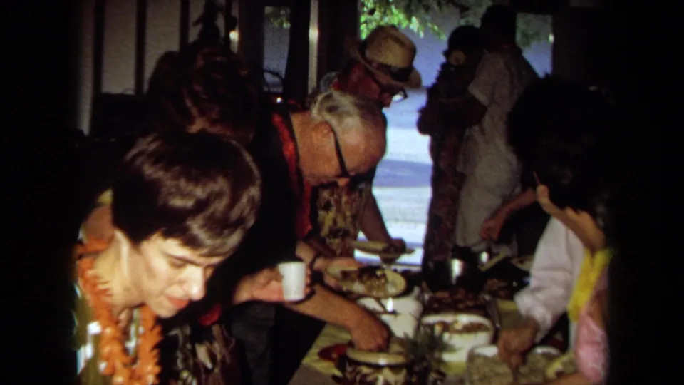 1971 California party frame showing guests in leis and patterned clothing serving themselves from a crowded buffet table.