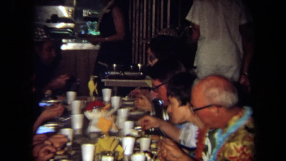 1971 California home-movie frame of adults seated closely at a party table eating and talking near cups and decorations.