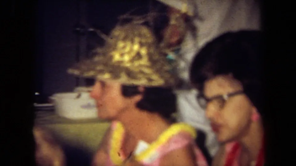 Soft-focus 1971 California frame of guests in leis near serving dishes, including one person wearing a straw hat.