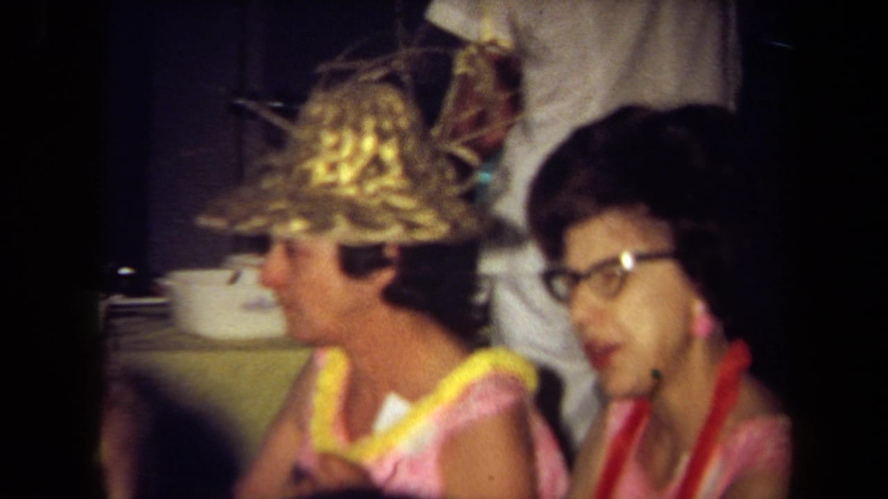 1971 California 8mm frame of two seated women at a busy indoor party or restaurant, with leis, a straw hat, and soft handheld focus.