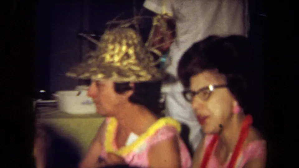 1971 California 8mm frame of two seated women at a busy indoor party or restaurant, with leis, a straw hat, and soft handheld focus.