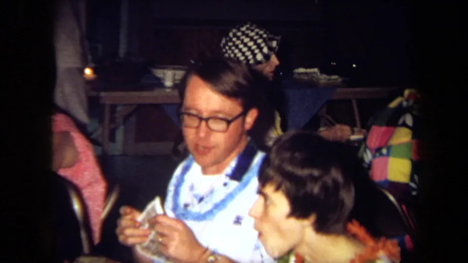 1971 California 8mm party frame showing a man with glasses and a blue lei at a dining table, with other guests partly visible around him.