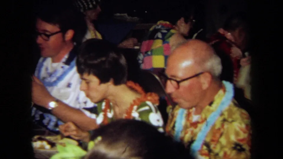 1971 California home-movie frame of adults seated around a party dining table, with aloha-style clothing, leis, plates, and low interior light.