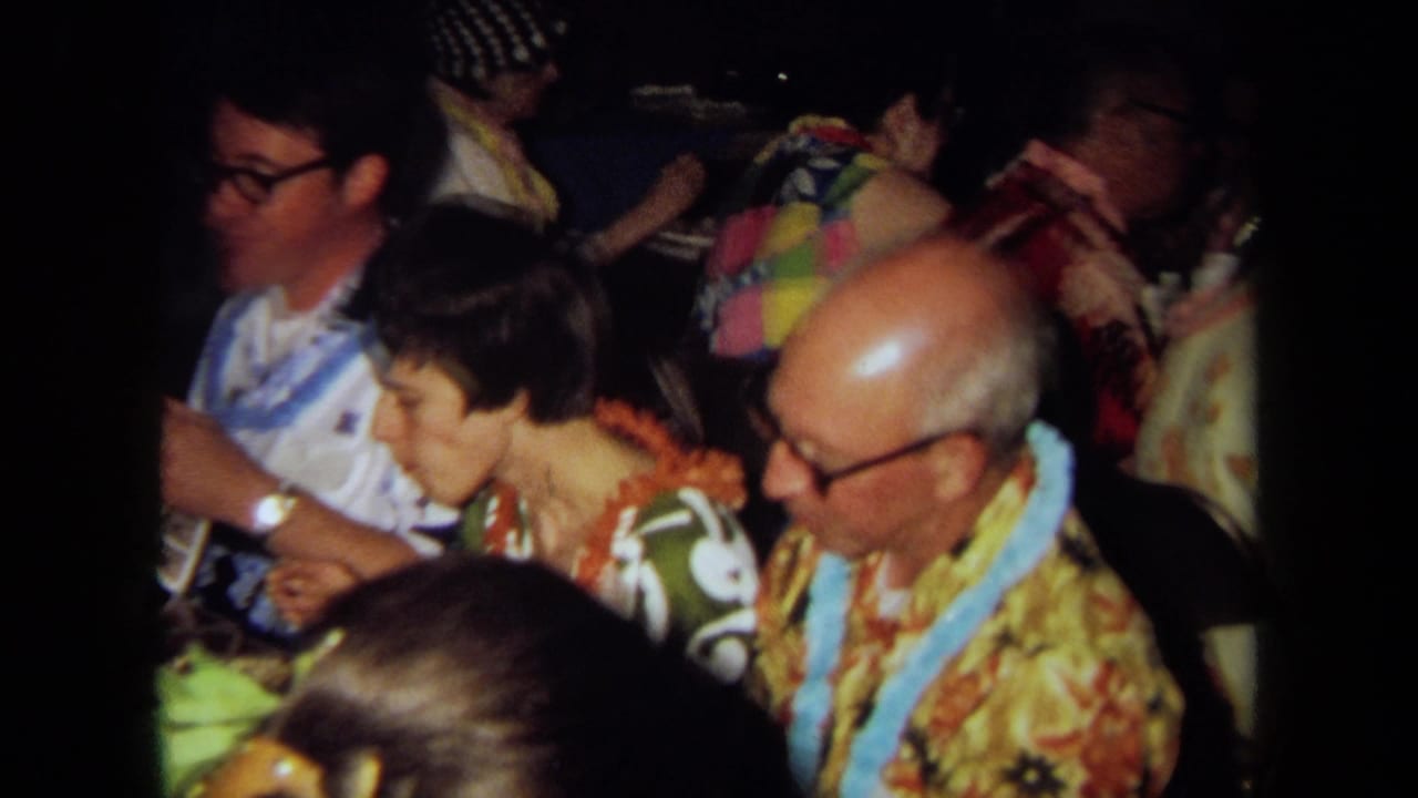 1971 California home-movie frame showing adults in aloha-style clothing and leis gathered closely around an indoor table.
