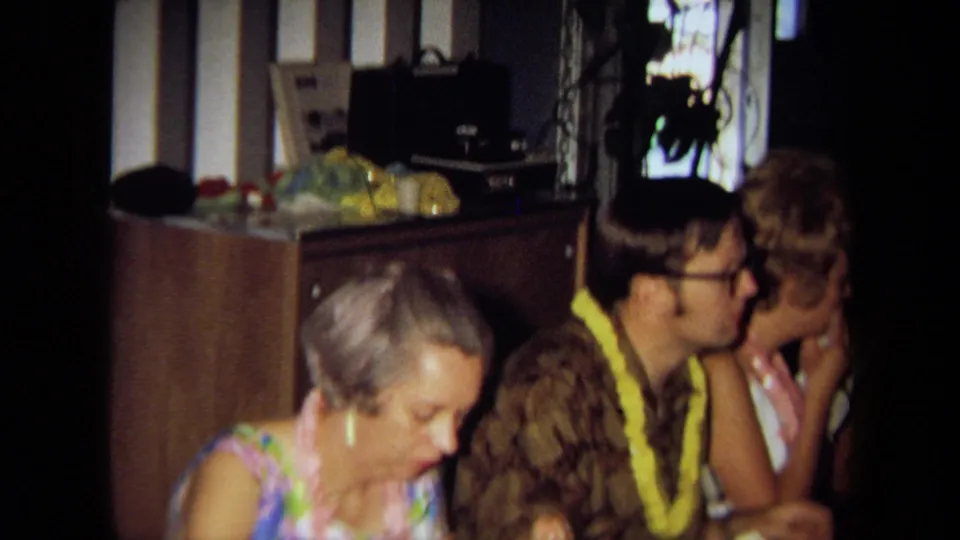 Adults at a 1971 California indoor party sit near a sideboard, window, and audio equipment while wearing floral leis.