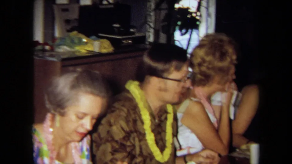 A 1971 California home-movie frame shows party guests in leis seated close together at a dining table inside a private home.