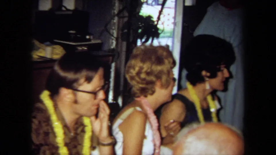 Party guests in a 1971 California private interior sit by a window, with leis, eyeglasses, and warm 8mm color visible.
