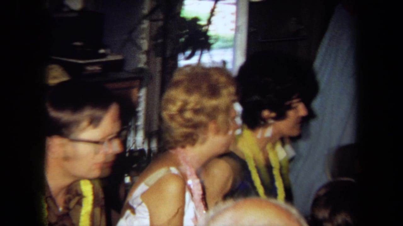 1971 California home-movie frame showing seated adults in conversation at an indoor party, with leis and a domestic window visible.