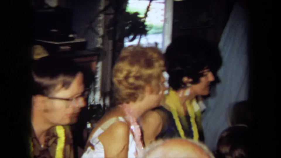 1971 California home-movie frame showing seated adults in conversation at an indoor party, with leis and a domestic window visible.