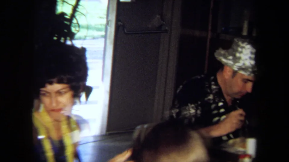 1971 California home-movie frame showing party guests seated by a table, including a man in a shiny silver novelty hat.