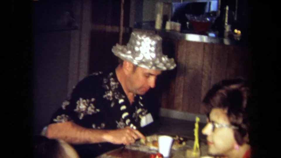 1971 California indoor party frame showing a seated man in a shiny silver hat eating beside another guest in a lei.