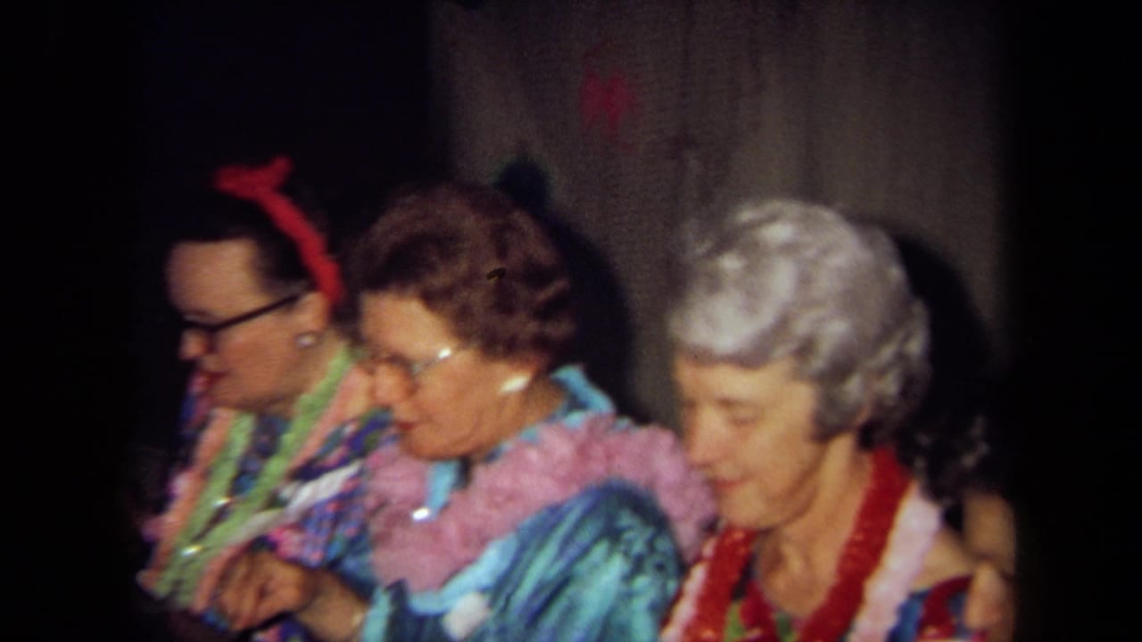 Three adults wearing leis sit shoulder to shoulder in a wood-paneled interior during a 1971 California home-movie party.