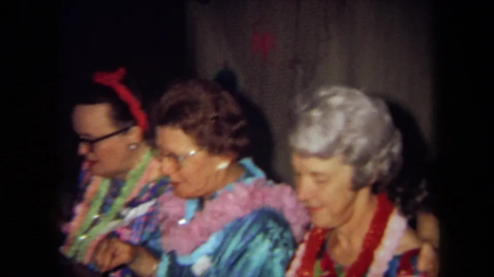 Three adults wearing leis sit shoulder to shoulder in a wood-paneled interior during a 1971 California home-movie party.