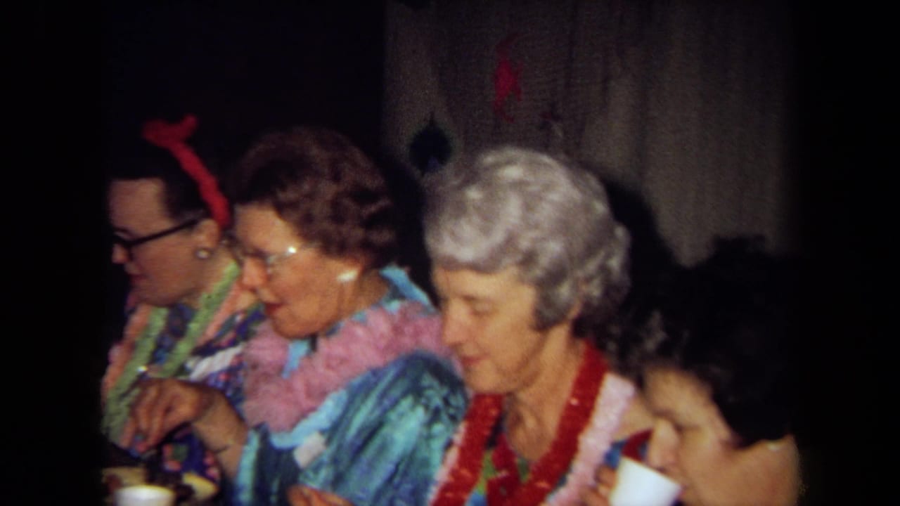 1971 California home-movie frame showing older party guests in aloha clothing and leis seated at a meal indoors.
