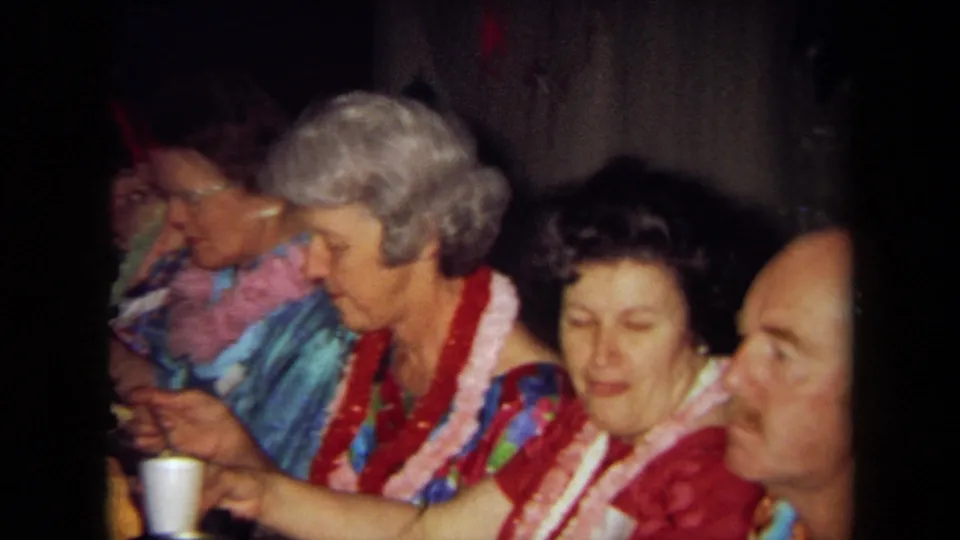 Older guests in leis and patterned shirts share food at a Hawaiian-themed party table in a California home-movie frame.