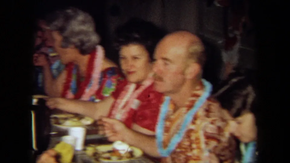 Home-movie frame of guests wearing leis at a crowded indoor meal, with plates, cups, and patterned clothing visible.