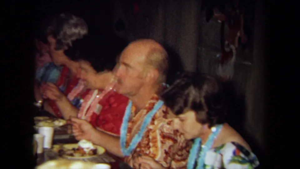 1971 California 8mm frame showing seated guests in leis eating at a Hawaiian-themed indoor gathering.