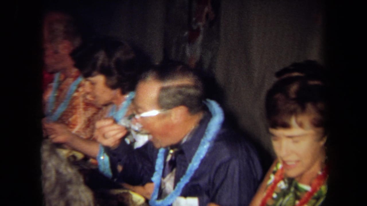 Adults wearing leis eat and talk at a crowded indoor table.